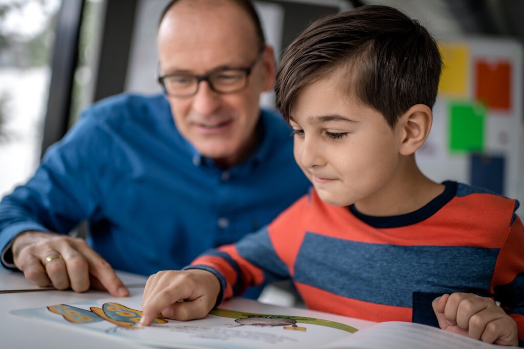 Foto von einem Mann und einem Jugen, die an einem Tisch sitzen und in einem Buch gemeinsam lesen.